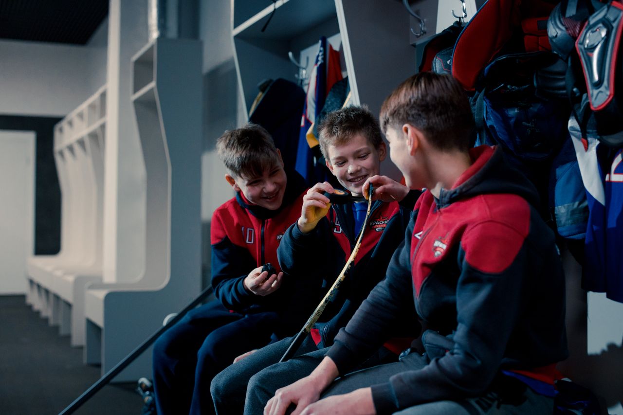 Three young boys in hockey gear taping sticks and laughing in a locker room.
