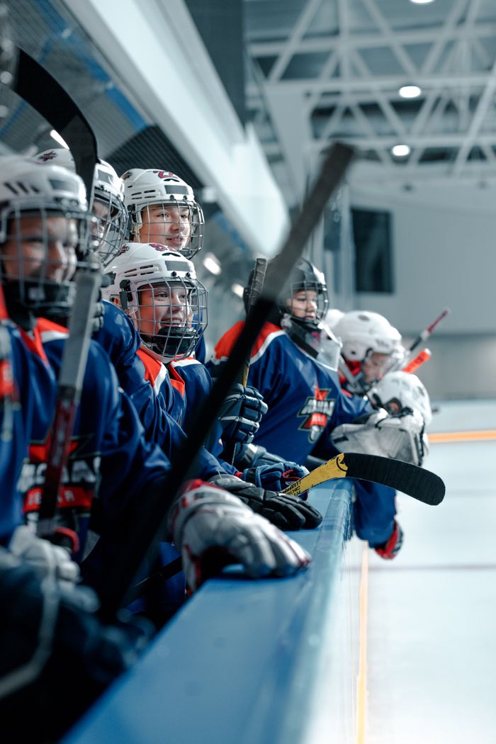 A youth hockey team with helmets and sticks waits on the bench at an indoor ice rink.